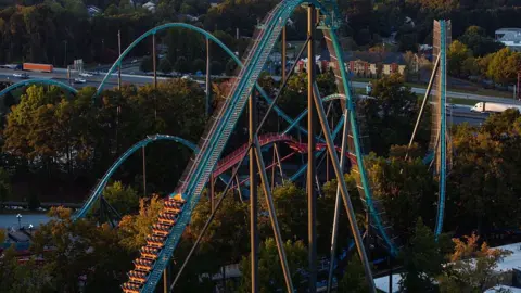 Getty Images Aerial view of large teal and orange rollercoaster, with train car stopped halfway up one of the coaster's peaks