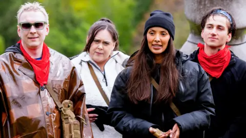 BBC/Studio Lambert/Euan Cherry Joe Lycett, Sharon Rooney, Maya Jama and Bash standing in a row outdoors with red neckerchiefs during a mission