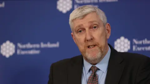 PA Media John O'Dowd. He has short white hair and beard, wearing a black blazer, light blue shirt and tie. Blue NI executive branding is behind him.
