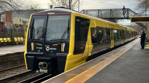 BBC News/Andy Gill A new Merseyrail train stops at a station platform. There's a man standing beside it, and a man on a bridge over the tracks.