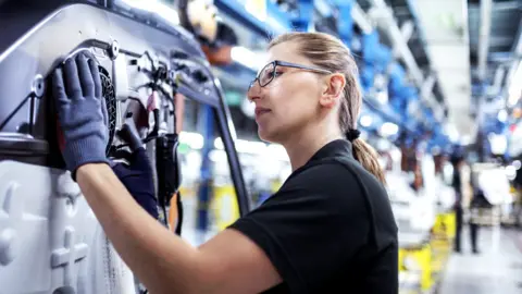 Getty Images A woman in a black T-shirt and wearing glasses and protective gloves works on a machine in a factory