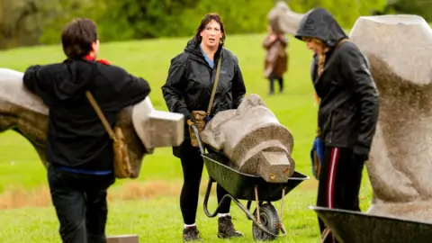 BBC/Studio Lambert/Euan Cherry Miranda in a raincoat pushing a wheelbarrow with a stone head across the grass, in between two other celebrities and parts of statues
