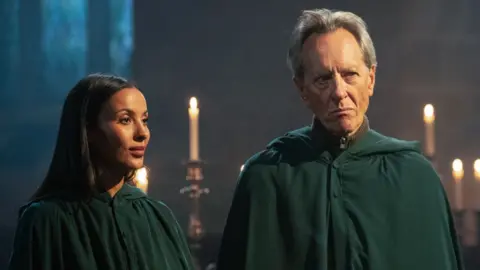 BBC Maya Jama and Richard E Grant wearing the traitors' green cloak