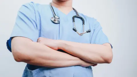 Getty Images A person wearing medical scrubs has a stethoscope around their neck and is folding their arms.