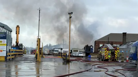 Thick grey smoke rising into the sky as fire fighters feed a hose from the back of a fire engine