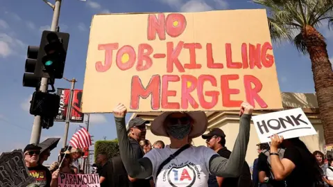Getty Images Protester holds a sign reading "No job-killing merger" in front of a crowd outside Paramount Studios in Los Angeles in September, 2026.